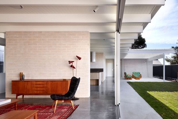 A refined modern living room opening onto a landscaped courtyard through full-height glazed doors. The room features a warm timber sideboard, black lounge chair, red patterned rug, pale brick feature wall, polished grey concrete floor, and exposed white ceiling beams. A covered outdoor walkway and lawn are visible beyond the glazing.