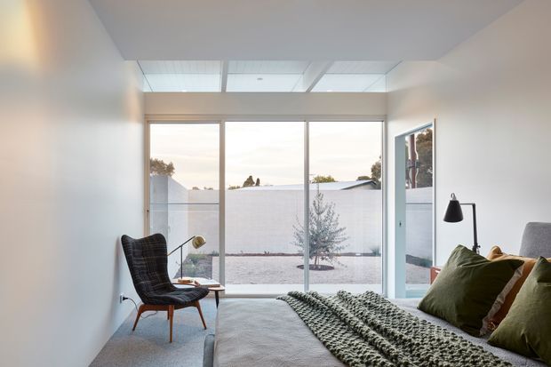 Bright, minimalist bedroom with a low-profile bed, textured green throw, olive cushions, a dark lounge chair, and a slim floor lamp. Large floor-to-ceiling glazing and overhead skylights provide views into a private courtyard and abundant natural light. The room has a restrained white and neutral palette with subtle mid-century-inspired furniture.
