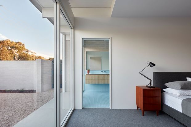 A bright contemporary bedroom viewed beside full-height glazing, with a grey upholstered bed, white bedding, dark grey carpet, and a mid-century-style timber bedside cabinet with a black angled lamp. Through the open doorway is a compact bathroom with blue patterned floor tiles, a timber vanity, and pale blue wall surfaces. The glazing opens toward a minimalist landscaped courtyard with a white masonry boundary wall.
