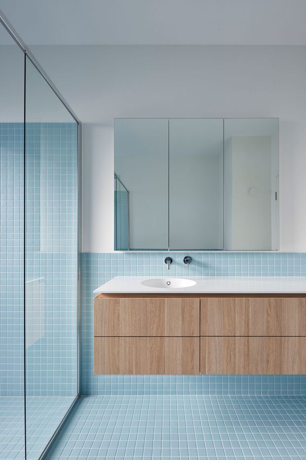 Bright minimalist bathroom featuring a frameless glass shower enclosure, pale blue small-format square tiles covering the shower walls and floor, a floating natural wood vanity with integrated white basin, wall-mounted chrome tapware, and a large mirrored medicine cabinet.
