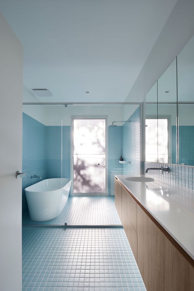 A bright contemporary bathroom with turquoise-blue mosaic tile covering the walls and floor, a freestanding white bathtub, frameless glass shower enclosure, and a long timber-look vanity with a white integrated benchtop and oval basin. Large mirrors reflect the vanity and shower area, while frosted windows provide diffuse natural light. The space has clean lines and a calm, spa-like atmosphere.