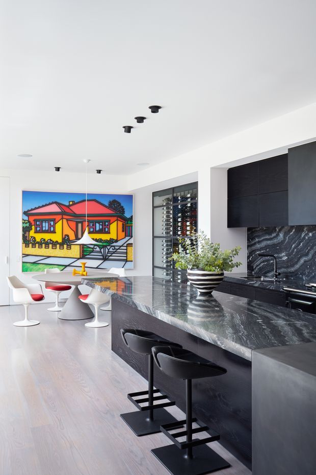 Bright, open-plan contemporary kitchen and dining area with a large dark stone island, black fixed cabinetry, dramatic veined stone splashback, integrated wine display, and sculptural black bar stools. A colourful framed artwork and distinctive white and red dining chairs add bold accents. The room has a clean white ceiling, pale timber flooring, and recessed and directional lighting.