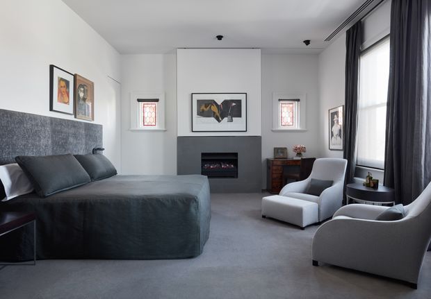 A refined bedroom with a large upholstered bed, grey carpet, dark charcoal curtains, and a matching textured charcoal feature wall behind the bed. The room includes a contemporary fireplace with a dark surround, two light armchairs by the window, a small side table, framed artwork, and compact timber storage furniture. Tall windows provide soft natural light, creating a calm, monochromatic and sophisticated atmosphere.