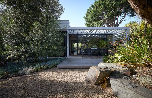 A modern single-storey house is viewed from a landscaped garden, with a covered outdoor terrace framed by slim dark metal posts and a translucent or slatted roof. Full-height glazing opens onto the terrace, revealing outdoor seating and the interior beyond. Gravel paving, large natural timber or stone-like garden edging, sculptural rocks, and established planting surround the structure.