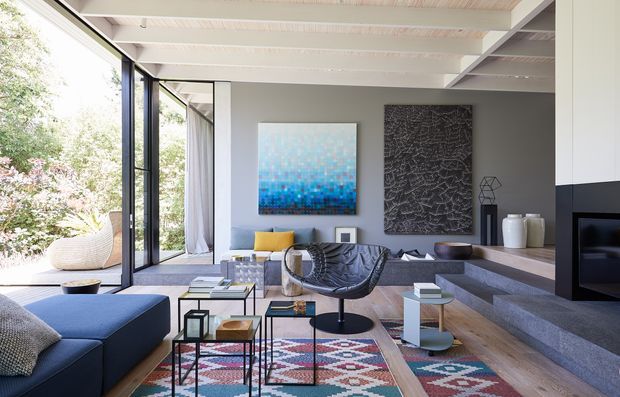 Bright contemporary living room with an open-plan arrangement, floor-to-ceiling black-framed glazing opening to a leafy garden, exposed white-painted ceiling beams, pale timber flooring, a large dark sectional sofa, sculptural lounge chair, layered geometric rugs, and low occasional tables. A stepped stone fireplace wall and abstract artwork add strong architectural and decorative features.