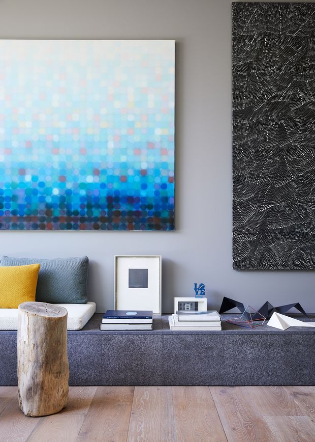 A contemporary living room vignette with a low, dark grey built-in or upholstered console beneath two large abstract artworks. The room features a grey sofa with a mustard yellow cushion, a natural timber stump coffee table, layered books and framed objects, geometric decorative sculptures, and pale timber flooring. The palette is cool grey and blue with warm yellow and natural wood accents.