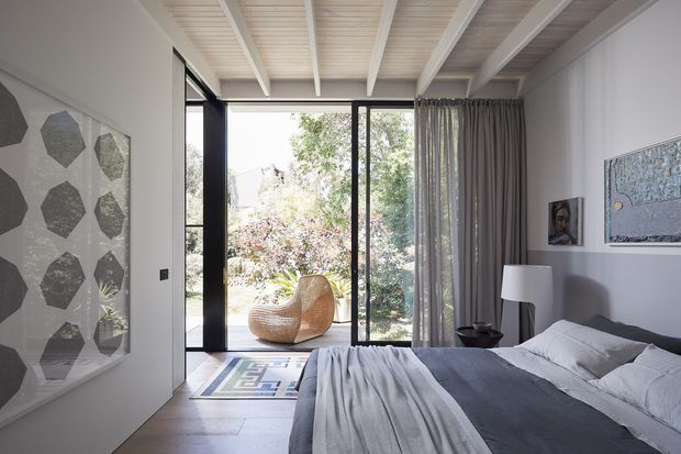Bright contemporary bedroom with a low-profile bed dressed in layered grey bedding, a white bedside lamp, framed artwork, and a sculptural woven lounge chair. Full-height black-framed glazing opens to a leafy garden, while sheer grey curtains provide privacy. The room features a distinctive white-painted exposed-beam ceiling and pale timber flooring.
