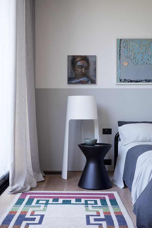 A softly lit contemporary bedroom corner with a black metal-framed bed partially visible at right, layered white and grey bedding, a tall white floor lamp, and a sculptural black side table. Full-height sheer and linen curtains frame a window on the left. The pale grey wall displays two abstract artworks, while a colourful geometric rug sits over a warm timber floor.