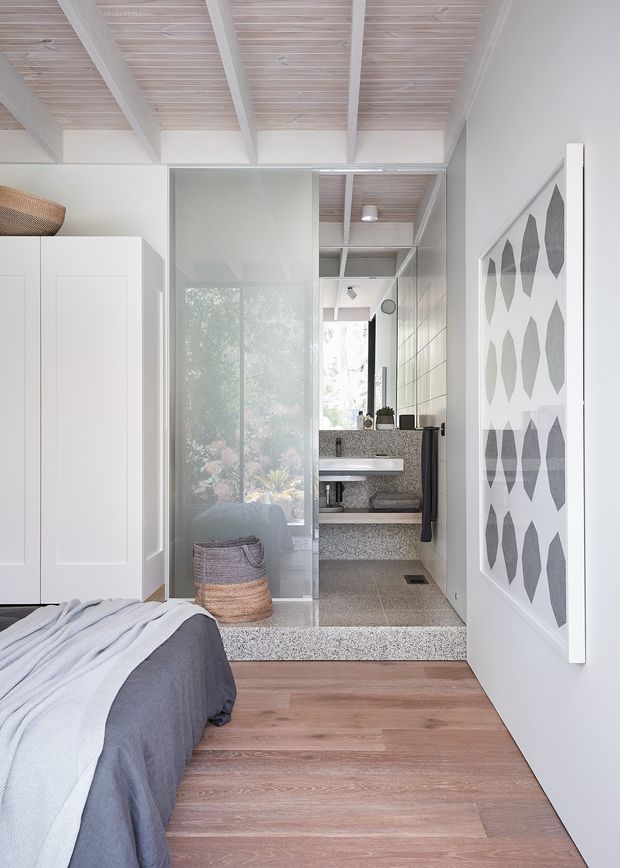 A bright contemporary bedroom with a partially visible bed, white built-in wardrobe, pale timber floor, and exposed white-painted ceiling beams with timber infill. A translucent glass screen separates the bedroom from a compact bathroom visible beyond, featuring a wall-mounted basin, terrazzo-like benchtop and tiled surfaces. A geometric monochrome artwork hangs on the right wall, while a woven basket adds texture.
