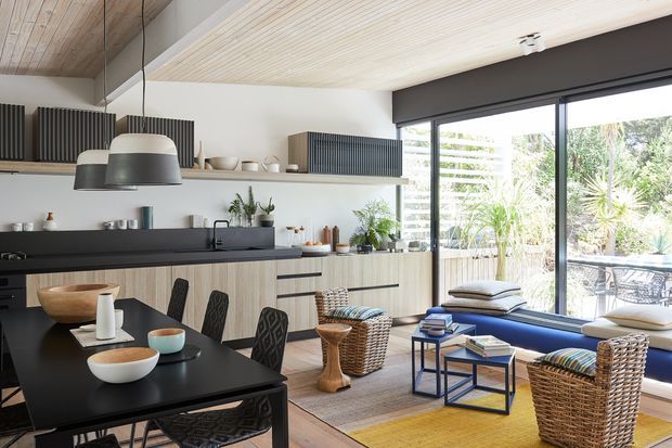 Open-plan contemporary kitchen and dining-living space with a black dining table in the foreground, pale wood kitchen cabinetry, dark benchtops, open shelving, pendant lighting, and a blue built-in window seat. Woven wicker lounge chairs, layered rugs, books, and plants create a warm, relaxed interior. Large glazing opens the room visually to a planted outdoor area.