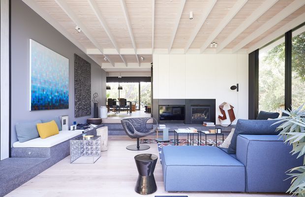 Bright, refined contemporary living room with a large blue sectional sofa, pale upholstered bench seating, glass and metal coffee tables, a central fireplace wall, expansive black-framed glazing, and a distinctive white exposed-beam ceiling. Blue artwork and a yellow cushion provide colour accents.