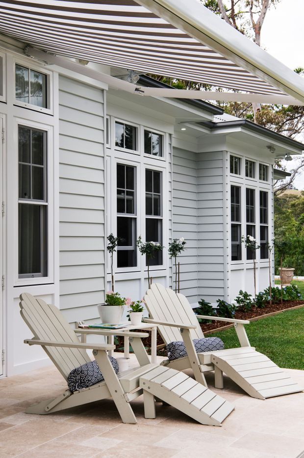 A bright exterior patio beside a white weatherboard-clad home, featuring tall multi-pane windows, a striped retractable pergola or awning, pale tiled paving, and two light-coloured reclining outdoor lounge chairs with cushions and a small table. Garden beds and established trees surround the house.