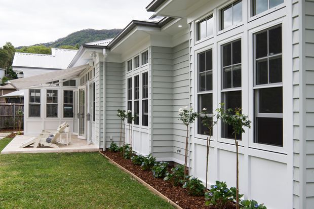 Exterior view of a light-coloured weatherboard residential extension with extensive white-framed windows and glazed doors. A covered outdoor sitting area connects to the main facade, with landscaped garden beds and lawn in the foreground and a wooded hill in the background.