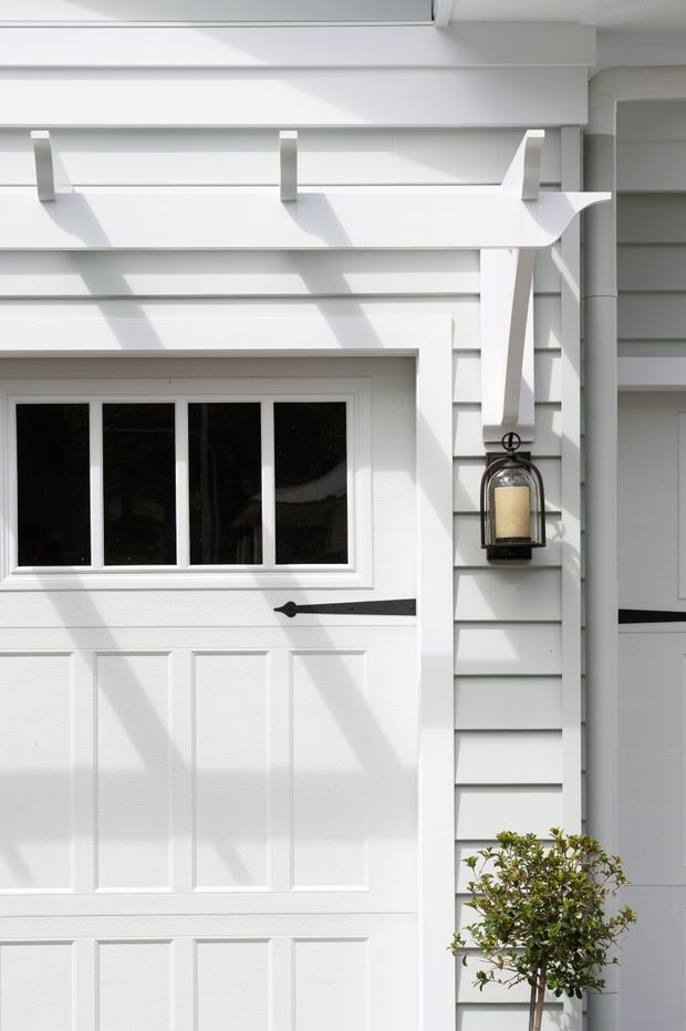 Close-up exterior view of a bright white residential facade featuring a paneled garage door with multi-pane window, horizontal weatherboard cladding, a projecting white awning, black decorative hardware, and a wall-mounted lantern. A small clipped shrub is visible at the base.