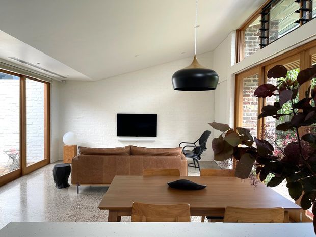 Open-plan living and dining area with a warm timber-framed glazed wall, white-painted brick feature wall, polished light terrazzo floor, and a mezzanine-level balustrade. A brown sectional sofa faces a wall-mounted television, while a timber dining table and chairs occupy the foreground. A large black pendant light, contemporary furnishings, and indoor planting complete the bright, relaxed interior.