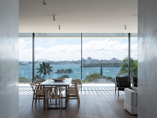 A bright, minimalist dining area opening onto expansive floor-to-ceiling glazing with a panoramic waterfront and city skyline view. A timber dining table with matching chairs occupies the foreground, with a dark lounge chair and part of a white sofa at right. Pale walls and flooring, slim dark window frames, recessed ceiling lighting, and strong striped sunlight create an airy contemporary interior.