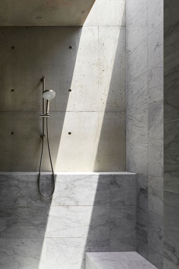 Minimalist bathroom shower area with a handheld and fixed shower fitting mounted on a raw concrete wall. Large white-grey marble-look tiles clad the lower walls and side wall, with a built-in tiled bench or ledge. Strong sunlight creates a dramatic diagonal beam across the surfaces, highlighting the restrained, monolithic material palette.