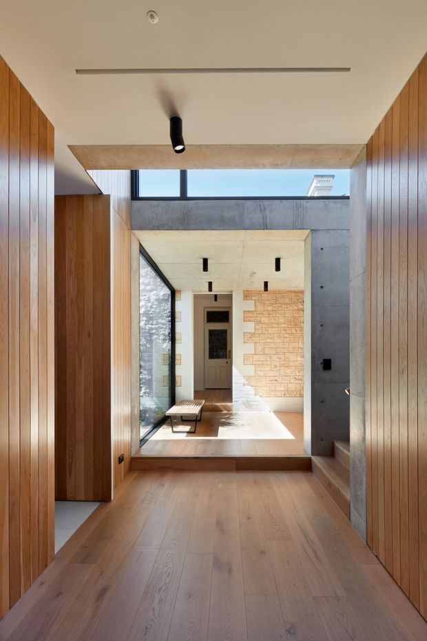 A sunlit residential hallway or entry passage with wide pale timber flooring, vertical warm timber wall panelling on both sides, exposed concrete beams and wall surfaces, and black recessed and cylindrical lighting. The corridor leads through a glazed opening and a small step into a brighter central space, with a textured pale brick or stone feature wall and a door visible beyond. The composition highlights layered thresholds, strong linear perspective, natural light, and a refined contemporary material palette.
