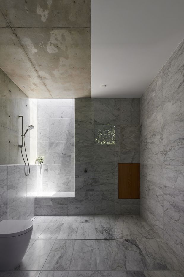 A minimalist contemporary bathroom with a walk-in shower, wall-mounted handheld shower, freestanding toilet, and a small timber wall cabinet. Large-format grey-white veined stone or marble tiles cover the floor and walls, contrasted by an exposed raw concrete ceiling and wall section. Natural light enters through a small window, creating a cool, calm and refined atmosphere.