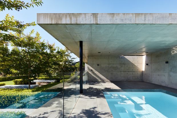 Exterior view of a contemporary concrete residence with a broad cantilevered flat roof and open ground-level terrace surrounding a swimming pool. Raw concrete walls and soffits create a strong minimalist architectural character, while frameless glass balustrades, landscaped planting, and crisp sunlight add contrast. The pool and covered outdoor area dominate the composition.