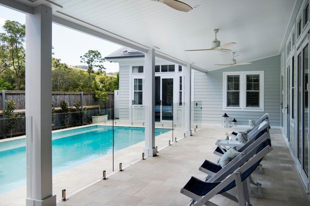 Bright covered outdoor terrace beside a swimming pool, featuring pale tiled paving, white-painted structural posts and ceiling, glass pool fencing, ceiling fans, and a light weatherboard-clad building with large glazed doors and windows. Several lounge chairs line the terrace, creating a relaxed poolside entertaining area.