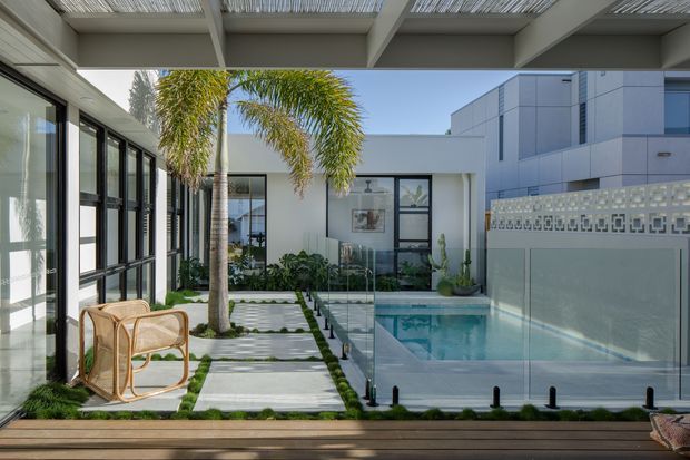 A bright contemporary courtyard pool area viewed from a covered outdoor walkway or terrace. The space features a compact rectangular swimming pool enclosed by clear glass balustrades, pale concrete stepping slabs separated by narrow strips of grass, tropical palms and planted beds, black-framed glazing, and a white rendered building with a decorative breeze-block privacy screen. A timber-toned deck and woven chair occupy the foreground.