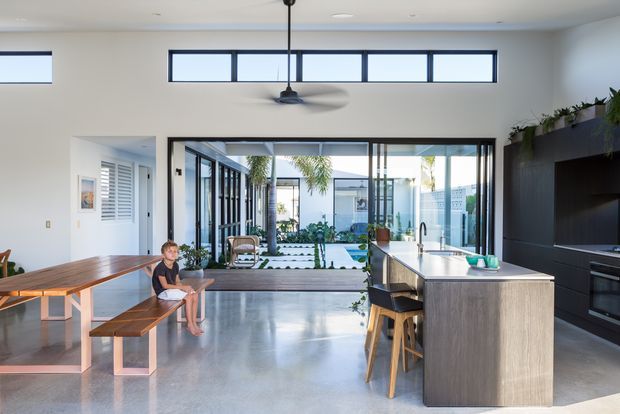 A bright, open-plan contemporary kitchen and dining area with polished concrete flooring, a large wood-look island with a light benchtop, dark built-in cabinetry, and timber dining furniture. Full-height glazed doors open toward a landscaped courtyard and pool area, while high-level clerestory windows and a central ceiling fan enhance the airy feel.