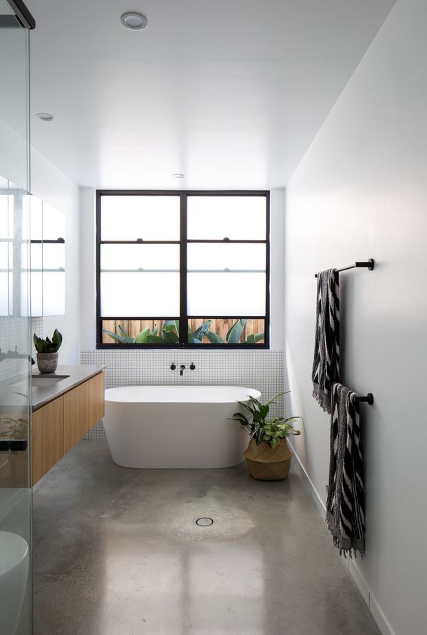 Bright contemporary bathroom with a freestanding white bathtub beneath a large black-framed window, a wall-mounted timber vanity with integrated basin, glass shower enclosure, small mosaic-tile wall section, polished concrete floor, and black towel rails. Potted plants and patterned towels add restrained natural and graphic accents.