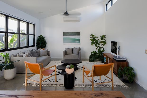 Bright contemporary living room with large black-framed windows, white walls, polished concrete flooring, a grey sectional sofa and armchairs, orange leather-and-timber lounge chairs, a round black coffee table, indoor plants, and a timber media console. A patterned rug anchors the seating area, while an exposed pendant light and split-system air conditioner are visible above.