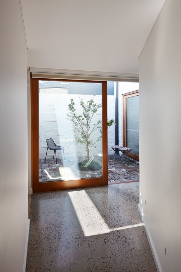 A narrow, bright interior passage leading to a large timber-framed glazed door opening onto a compact brick-paved courtyard. The corridor has white walls and ceiling, a polished speckled terrazzo or aggregate floor, and strong rectangular sunlight cast across the floor. Outside, a small tree, dark outdoor chair, and metal side tables are visible against pale painted masonry walls. The composition feels clean, minimalist, and connected to a private courtyard.