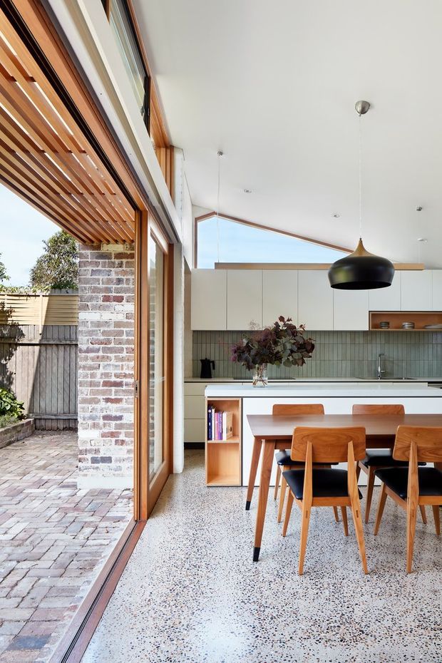 A bright, open-plan kitchen and dining area with large timber-framed glazed doors opening to a paved courtyard. The room features white flat-front cabinetry, a muted green-grey tiled splashback, a timber dining table and chairs, a large black pendant light, and a terrazzo floor. Exposed brickwork and timber slatted shading add warmth and texture.
