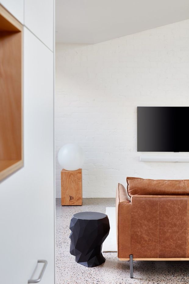 Bright modern living room viewed from an adjacent space, featuring a white-painted brick feature wall, wall-mounted television, brown leather sofa, white shelving and cabinetry, a faceted black side table, a spherical lamp on a timber pedestal, and a speckled terrazzo-style floor.