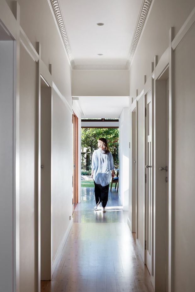 A bright, narrow residential hallway lined with multiple white internal doors and simple white trim. The corridor has a polished light timber floor, a high decorative ceiling with ornate linear moulding and recessed downlights, and leads through an open doorway to a sunlit living or outdoor area. A person is walking away from the camera near the far end.