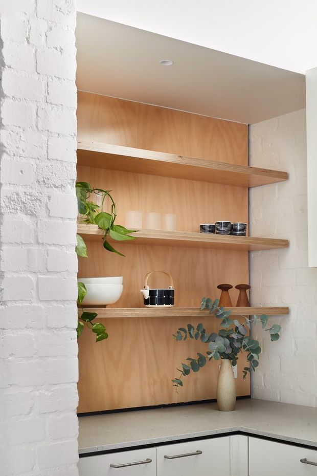 Bright contemporary kitchen detail featuring an open timber-lined shelving niche set into white-painted brick walls. Three timber shelves display crockery, cups, ceramics, plants and decorative objects above pale built-in cabinetry and a light benchtop. The composition has a warm natural timber backdrop contrasted with crisp white masonry and cabinetry.
