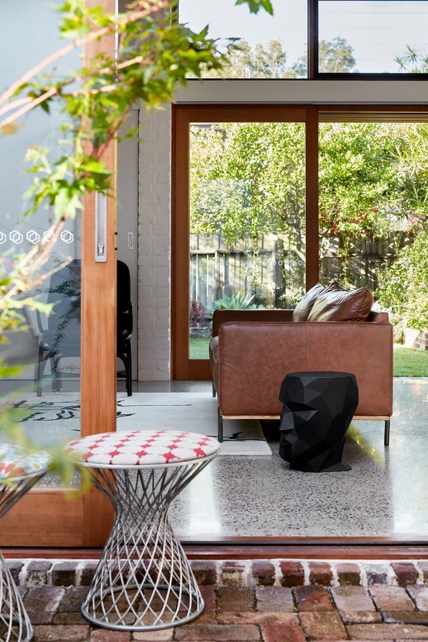 Bright contemporary living area viewed from an outdoor brick threshold, featuring full-height timber-framed glazing and doors, a white-painted brick wall, polished terrazzo flooring, and a brown leather armchair. A sculptural black side table and wire-base stool add graphic contrast, while greenery is visible through the glazing. The space has strong indoor-outdoor connection and abundant natural light.