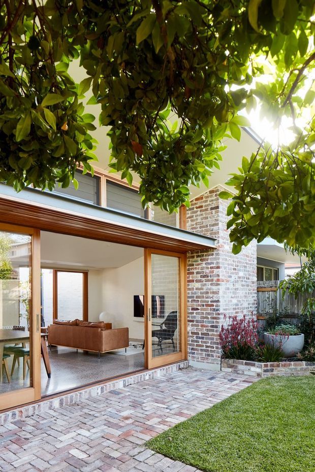 A bright residential courtyard viewed from the garden, showing a contemporary brick house with a timber-framed glazed living-room extension. Large sliding glass doors open onto a paved brick patio, with a sofa and television visible inside. Mature leafy planting forms a canopy overhead, while raised planters and a lawn surround the outdoor space.