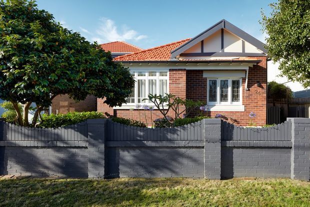 Street-facing view of a single-storey brick house with a prominent red tiled pitched roof, front gable, white-framed windows, and a dark painted masonry boundary wall. Mature trees and landscaped planting partially screen the facade.