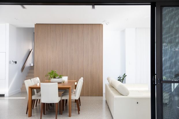 Bright contemporary open-plan interior showing a dining area and living room. A timber dining table with white upholstered chairs sits in front of a full-height vertical timber slat feature wall, with a staircase behind it. A white sofa, pale walls, terrazzo-like flooring, indoor plants, and large black-framed glazing complete the clean, light-filled space.