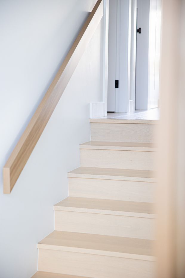 Bright minimalist stairway with pale timber treads and risers, a matching timber handrail, and white painted walls and door frames. The staircase rises toward a light-filled upper landing, creating a clean, airy interior.