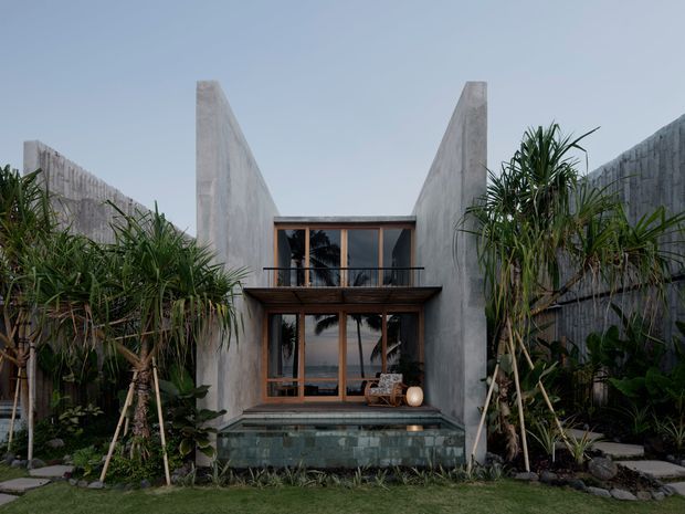 Front-facing exterior view of a compact, contemporary two-storey residence framed by lush tropical planting. Tall raw concrete side walls form a dramatic V-shaped roofline around dark timber-framed glazing, a small upper balcony, and a shallow tiled water feature or pool at ground level. Stepping stones and dense landscaping lead toward the facade.