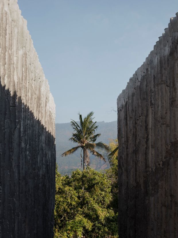 A narrow passage between two tall, heavily textured concrete or stone walls opens toward a tropical landscape. A palm tree and dense vegetation are visible in the gap, with hazy forested hills in the distance under a clear blue sky. The composition creates a dramatic framed view and strong contrast between the shaded walls and sunlit landscape.