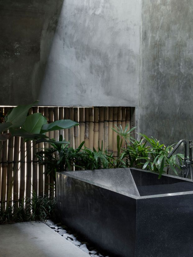 A moody, tropical bathroom courtyard featuring a large dark stone or concrete freestanding bathtub, exposed textured grey walls, bamboo screening, lush green plants, and a chrome wall-mounted tap. A smooth concrete floor and a strip of pale river stones complete the natural, spa-like setting.