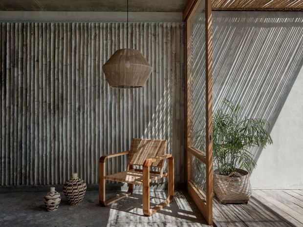 A warmly lit enclosed outdoor-style sitting area with a timber-framed glazed or screened wall, textured vertical feature wall, concrete floor, and a woven timber lounge chair. A large woven pendant hangs overhead, with ceramic vessels and a potted palm adding organic detail. Strong linear shadows fall across the floor and wall, creating a calm, tactile, indoor-outdoor atmosphere.