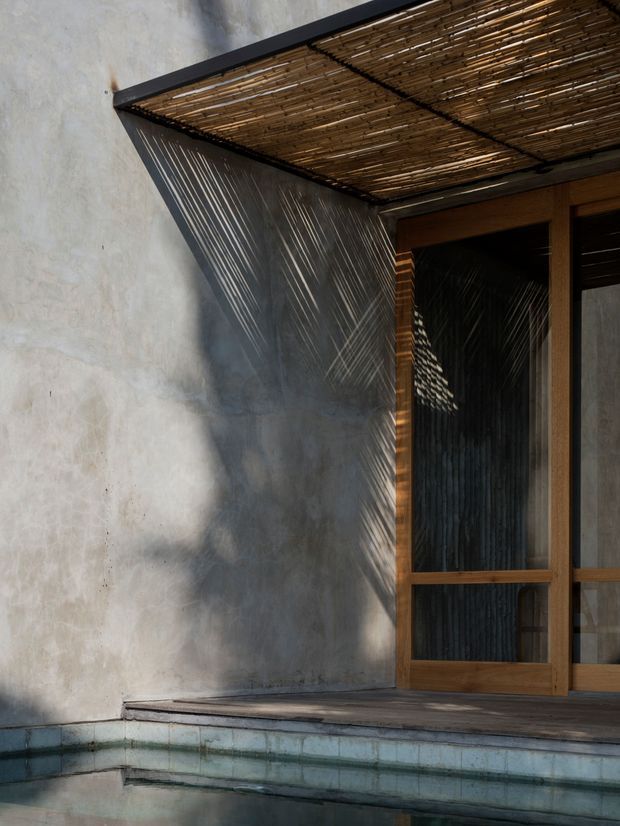 A close architectural detail of a shaded poolside threshold. A raw, pale rendered wall meets a timber-framed glazed doorway, with a projecting woven bamboo or reed canopy overhead casting strong linear shadows across the wall and glazing. Weathered timber decking leads to a small pool lined with pale blue-green tiles. The composition is minimalist and textural, with warm timber, rough plaster, patterned shade, and water at the foreground.