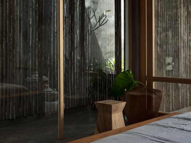 A moody bedroom interior viewed toward a dark glazed partition or window wall. Vertical textured translucent glass panels are framed with warm timber, with reflections revealing part of the room. A bed with light grey bedding occupies the foreground, alongside a sculptural timber stool and a large dark ceramic planter holding tropical foliage. The space has a restrained, natural-material palette and dramatic filtered light.