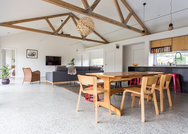 Open-plan living, dining and kitchen interior with exposed timber roof trusses, white walls, polished terrazzo-style flooring, and abundant natural light. A dark sofa faces a wall-mounted television, while a large timber dining table with timber chairs occupies the foreground. The kitchen includes timber cabinetry, a dark benchtop, open shelving, and red bar stools. A woven pendant light and indoor plants add warmth to the relaxed contemporary space.