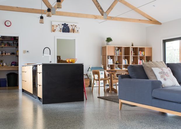 Bright open-plan interior combining a kitchen, dining area and living room. The room features a black kitchen island with timber cabinetry, polished concrete or terrazzo-style flooring, exposed timber roof trusses, open shelving, a timber dining table and a blue upholstered sofa. Natural light enters through a large window, creating a warm, airy and relaxed atmosphere.