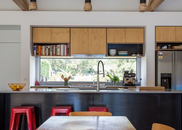 A contemporary kitchen viewed from the dining area, featuring a large dark island with integrated sink and tall gooseneck tap, warm timber overhead cabinetry, open shelving, stainless-steel appliances, and a wide horizontal window above the benchtop. Red and orange bar stools line the island, while exposed timber ceiling beams add warmth.