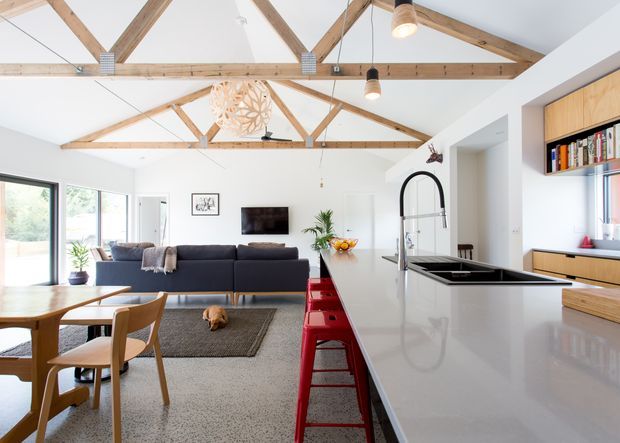 A bright open-plan kitchen, dining and living area beneath a high pitched ceiling with exposed timber trusses and beams. The kitchen features a large white island with an undermount sink, tall curved tap and red bar stools, alongside light timber cabinetry and open shelving. A timber dining table, dark sectional sofa, TV and large glazing create a contemporary, welcoming family space.