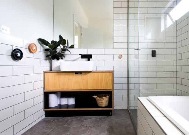Bright contemporary bathroom with white subway-tiled walls, a timber floating vanity with white basin and open black shelving, dark hexagonal floor tiles, a frameless glass shower enclosure, and a partially visible freestanding or built-in white bathtub. A large mirror and potted plant add light and softness.